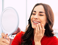 A young woman wearing a red blouse is smiling in the mirror while looking at her fully restored smile in Thorndale