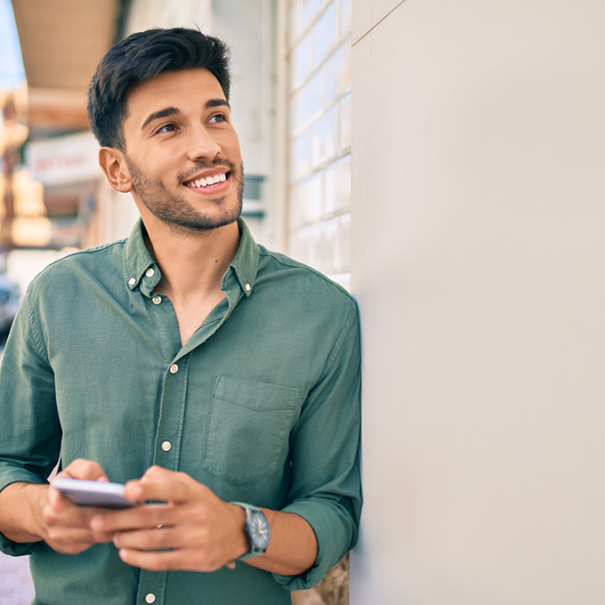 Smiling man leaning against building while checking his smartphone